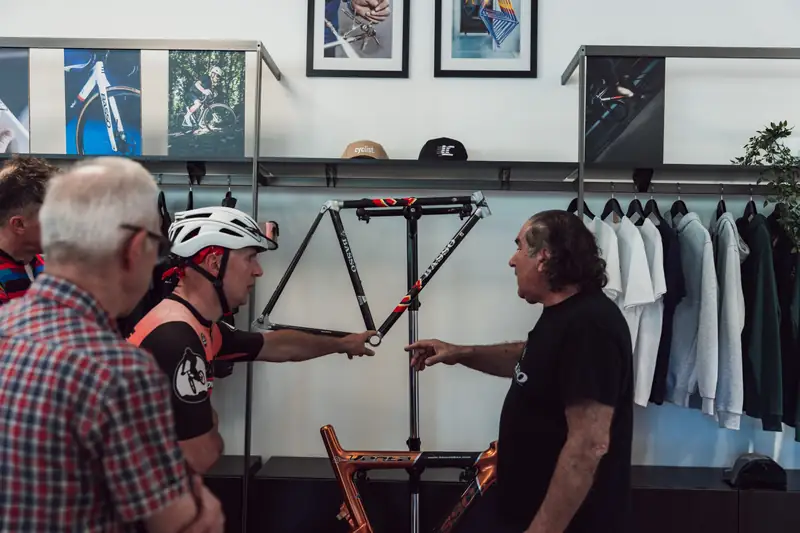 A man in a Basso T-shirt pointing at a bare Basso frame as he explains it to visiting riders in the showroom