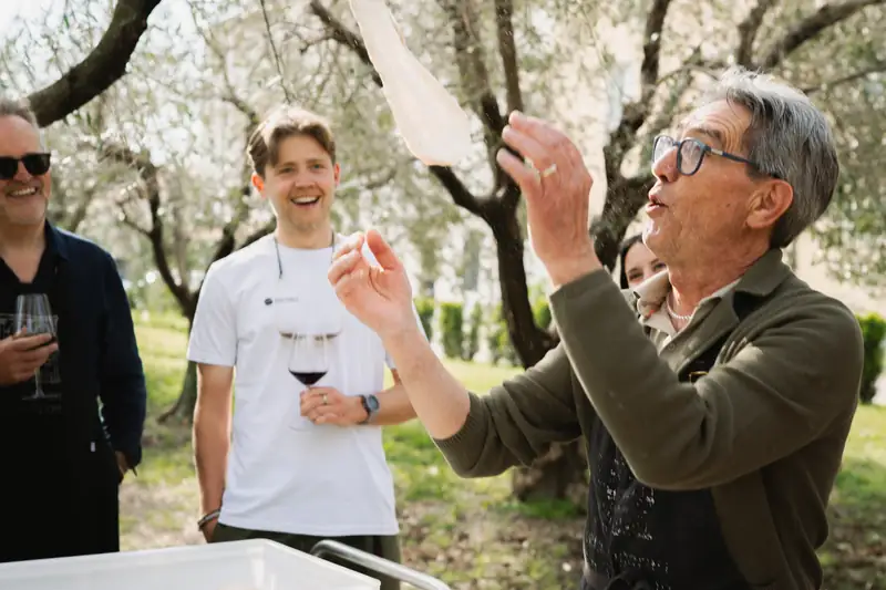 A host laughing as he lifts a sheet of dough among olive trees, guests behind him with glasses of wine