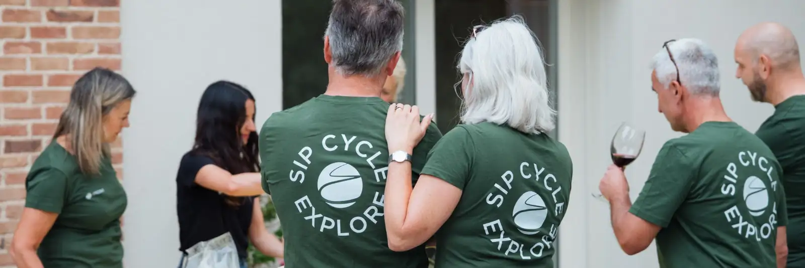 Guests in green Sip Cycle Explore shirts gathered round a table outside, one with a glass of red wine